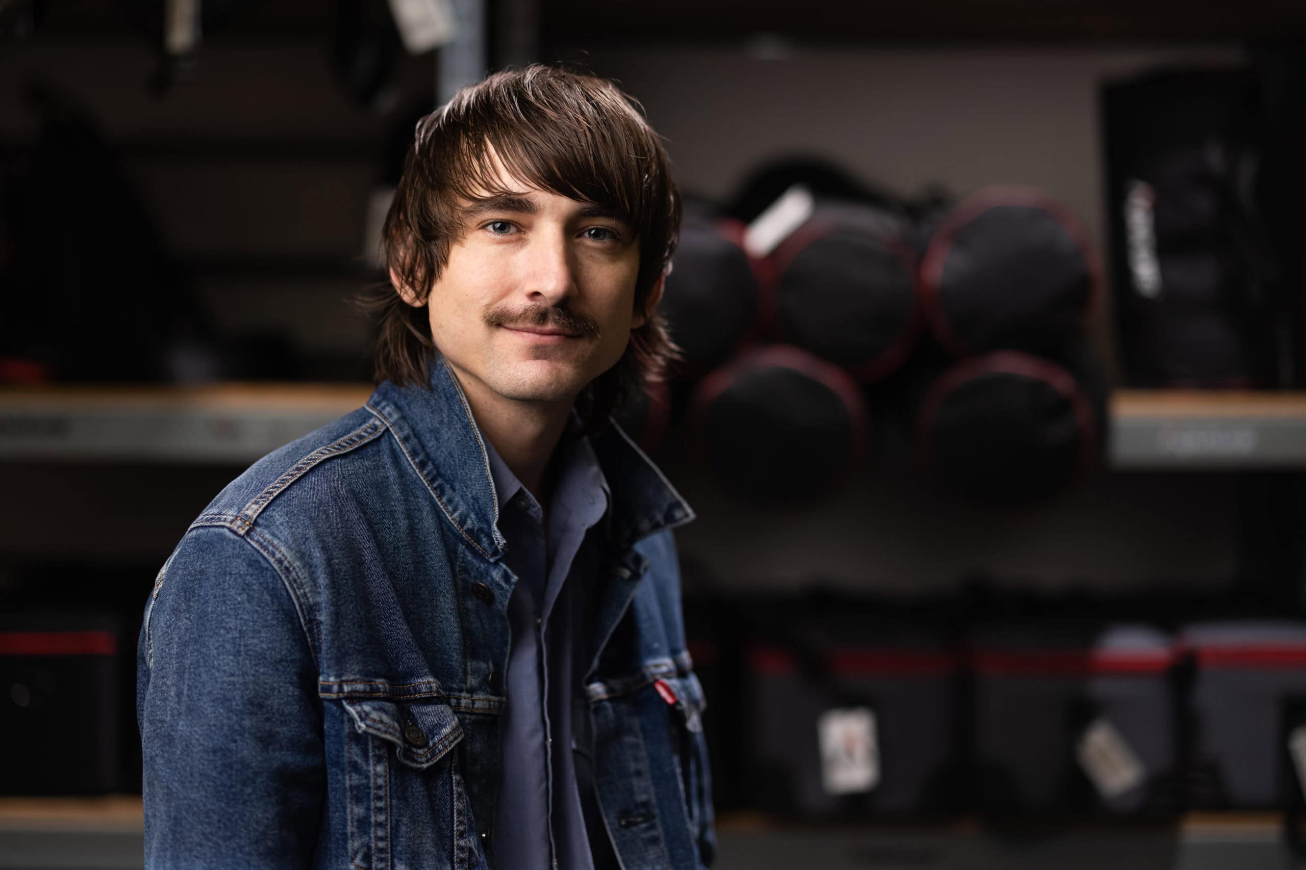 Photo of a man wearing a denim jacket in a camera rental store with camera gear in the background.
