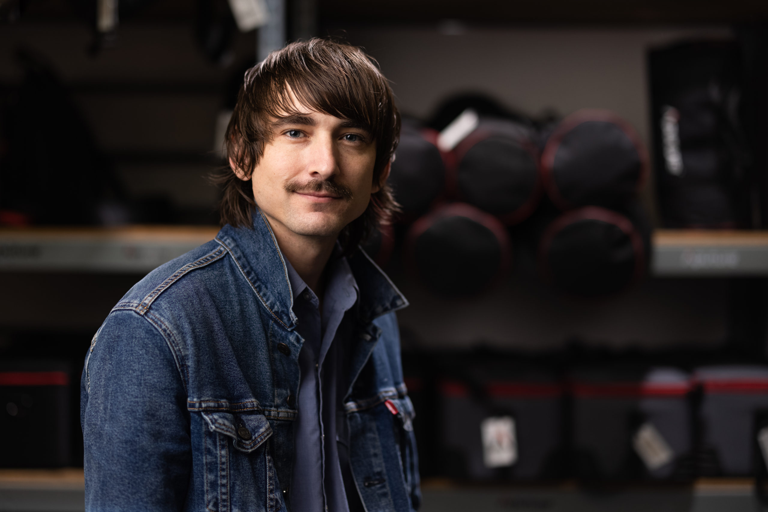 Photo of a man wearing a denim jacket in a camera rental store with camera gear in the background.